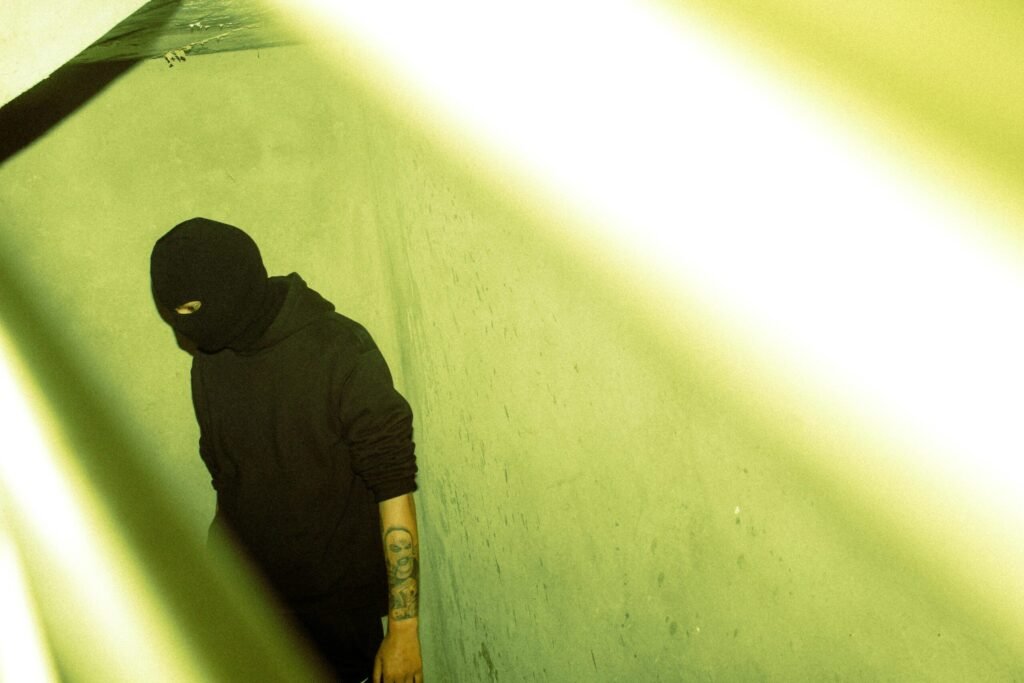 A person with a tattoo wearing a hoodie and mask stands in dim light, creating a mysterious atmosphere.